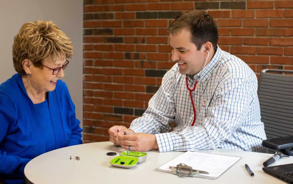 Dr. Brent Steele of The Hearing Guy consulting with an older woman patient and fitting a hearing aid at the brick-walled Asheville office