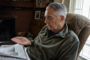 Older man holding a small receiver-in-canal hearing aid in his open palm, looking at it thoughtfully in his home