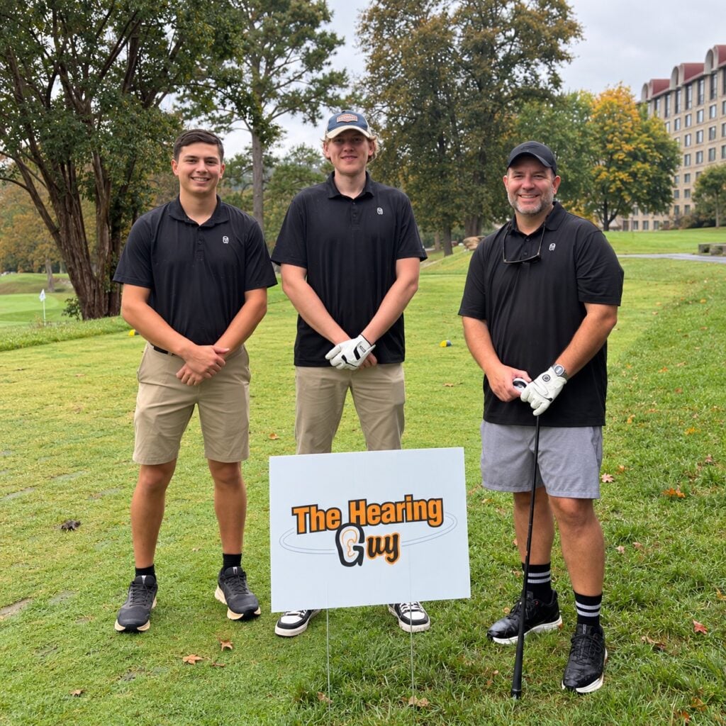 Three golfers beside The Hearing Guy sponsor sign at the Crush Cancer golf tournament at Grove Park Inn.