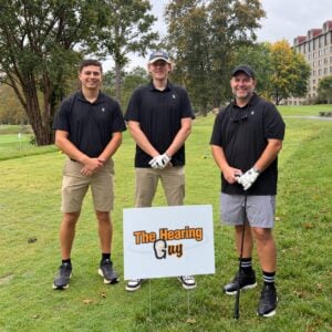 Three golfers beside The Hearing Guy sponsor sign at the Crush Cancer golf tournament at Grove Park Inn.